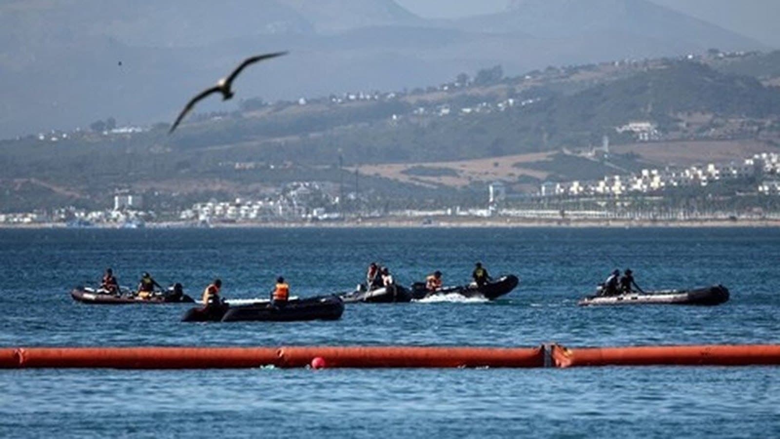 Inflatable boats working in the water off Ceuta beside a long orange floating barrier, with the Moroccan coast behind.