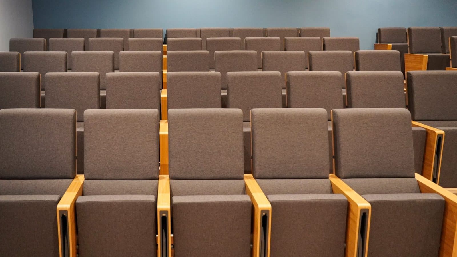 A small lecture hall with rows of empty, gray upholstered seats with wooden armrests facing the front. Photo d'archive.