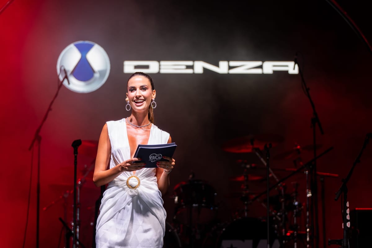 The host on stage in a white gown beneath the illuminated DENZA logo.