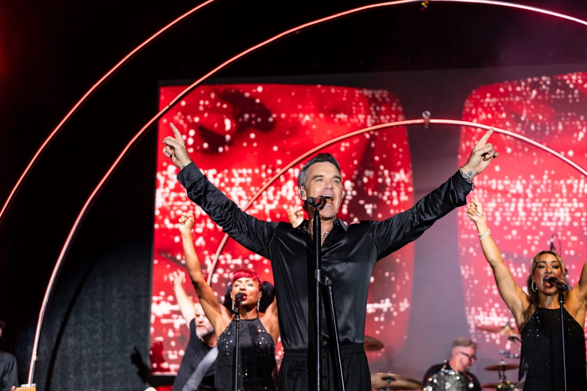 Robbie Williams on stage with both arms raised, his band behind him.